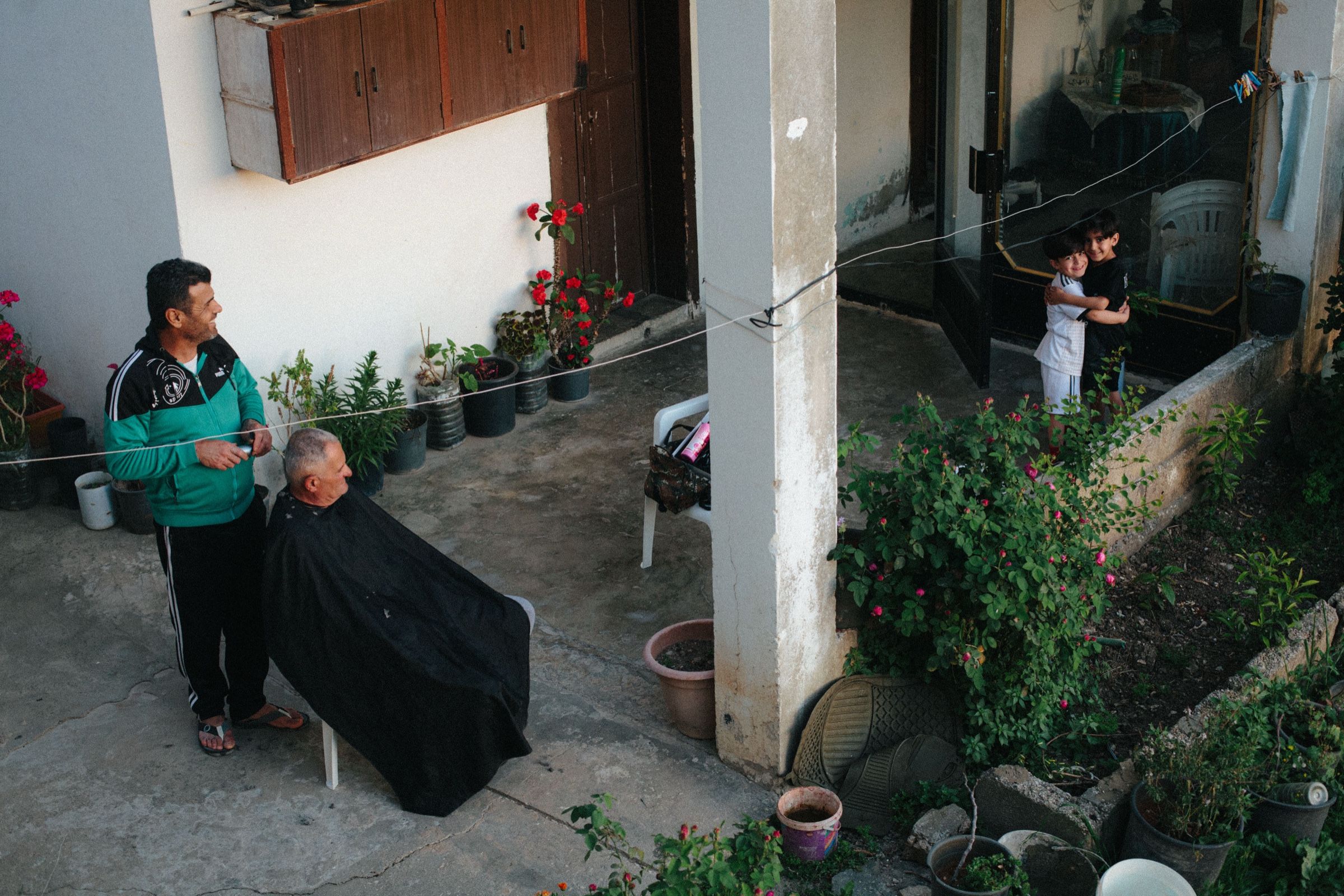 A man receives a haircut from a local barber in the courtyard garden of a modest home at dusk. Rows of potted plants line the perimeter. In the background, two children, distant relatives of the barber, embrace near the entrance, looking toward the camera. The town of Joun, Lebanon.