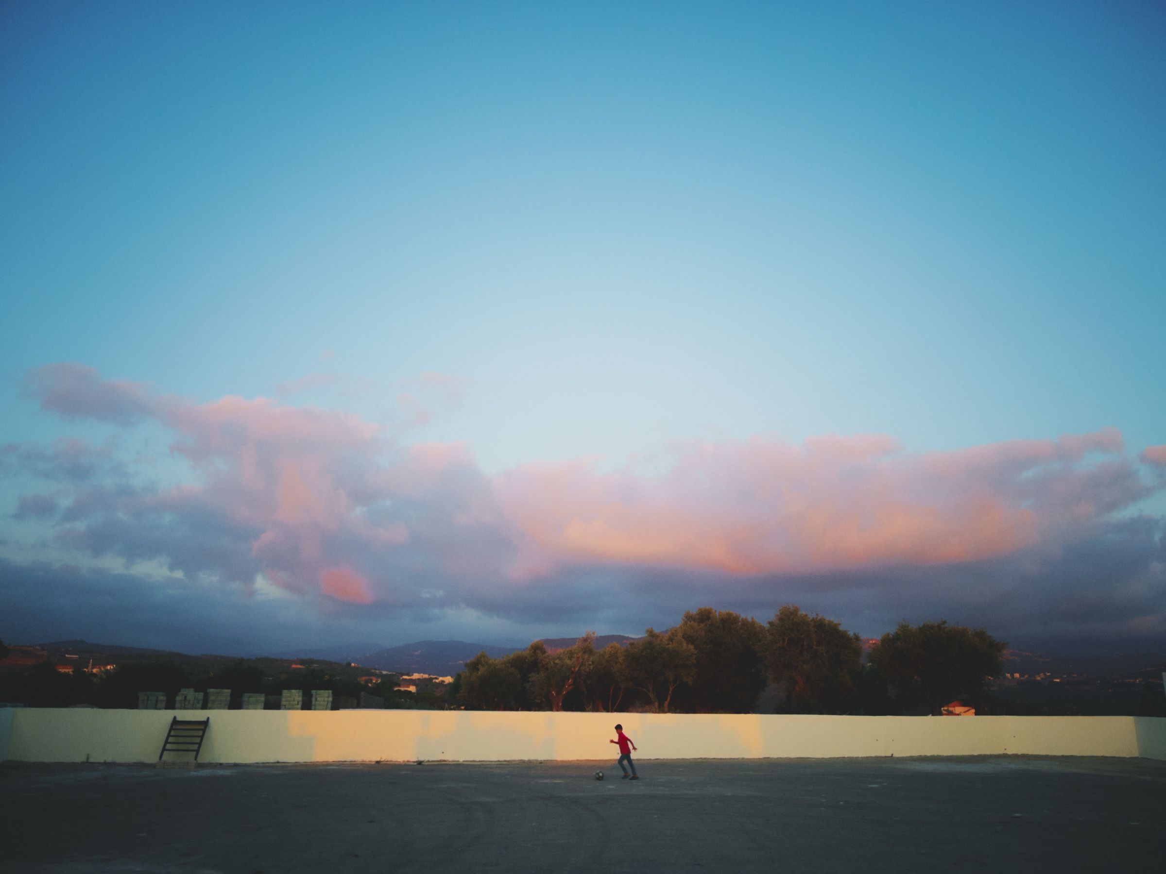 A boy plays alone with a soccer ball in an open space as clouds gather over the hills at dusk. The town of Joun, Lebanon, 2025.